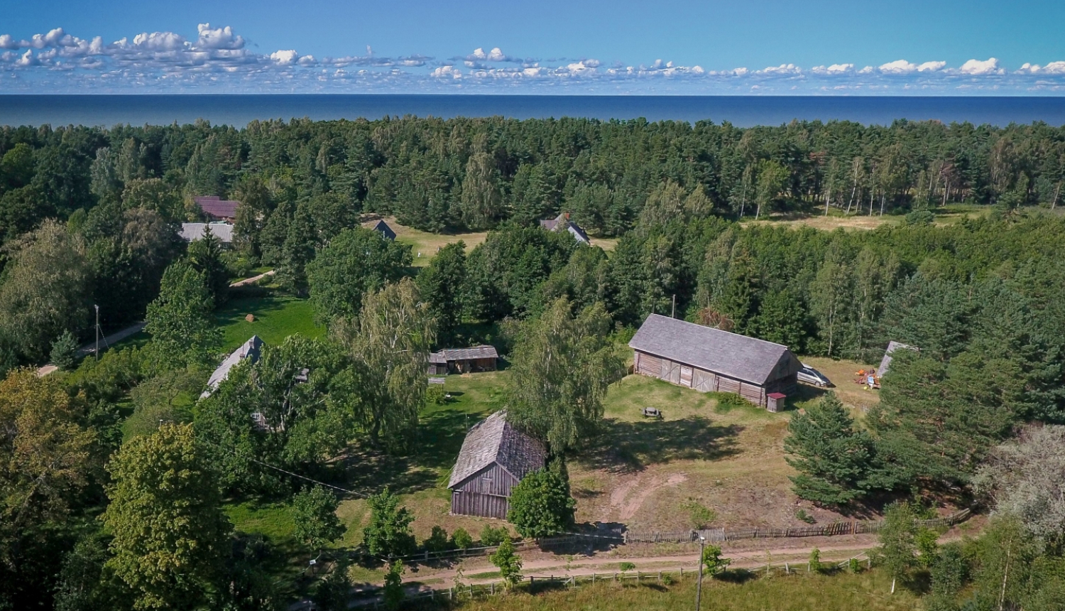 Attēlā redzams pilsētbūvniecības piemineklis - Košraga zvejniekciems. Pilsētbūvniecības piemineklis iekļauj plašāku cilvēka veidotu vidi, kā vecpilsētu vai ciemu. Attēlā redzams ar dronu no gaisa fotografētais Košraga ciems lībiešu krastā. Foto augšpusē pirmo ceturtdaļu aizņem tumši zilas vasaras debesis ar rotaļīgu baltu mākonīšu rindu virs horizonta, kam seko tumši zaļais priežu mežs. Priekšplānā var izšķirt kādu no ciema vēsturiskajām ēkām, starp kokiem šur tur manāmi zemi divslīpju jumti.