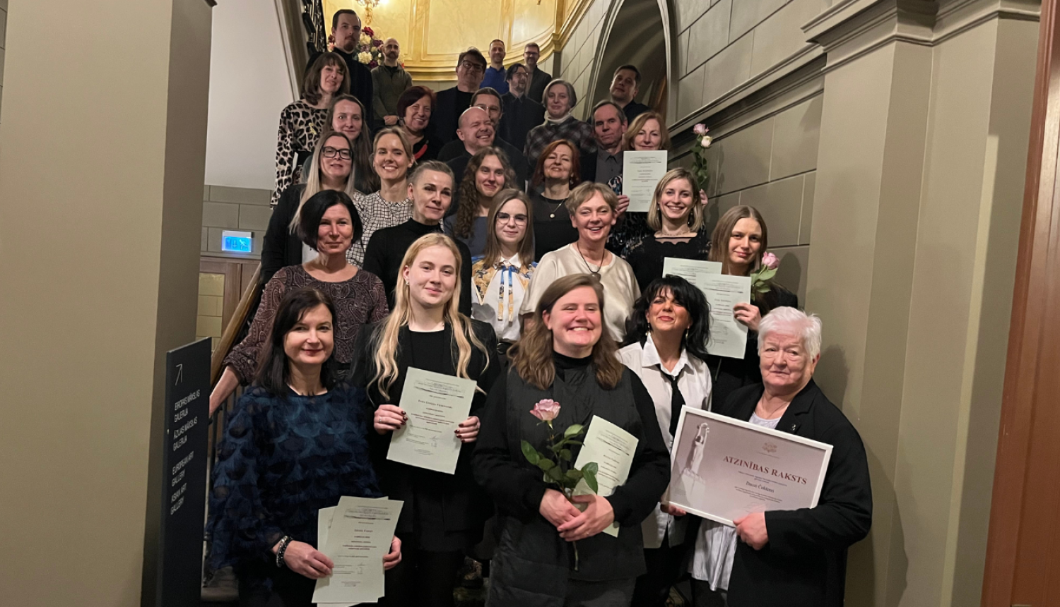 Grupas fotogrāfija uz kāpnēm, kurā redzami pasākuma dalībnieki un restauratori. Centrā, priekšplānā, redzamas sievietes ar diplomiem un viena sieviete tur Ministru kabineta Atzinības rakstu rāmītī.
