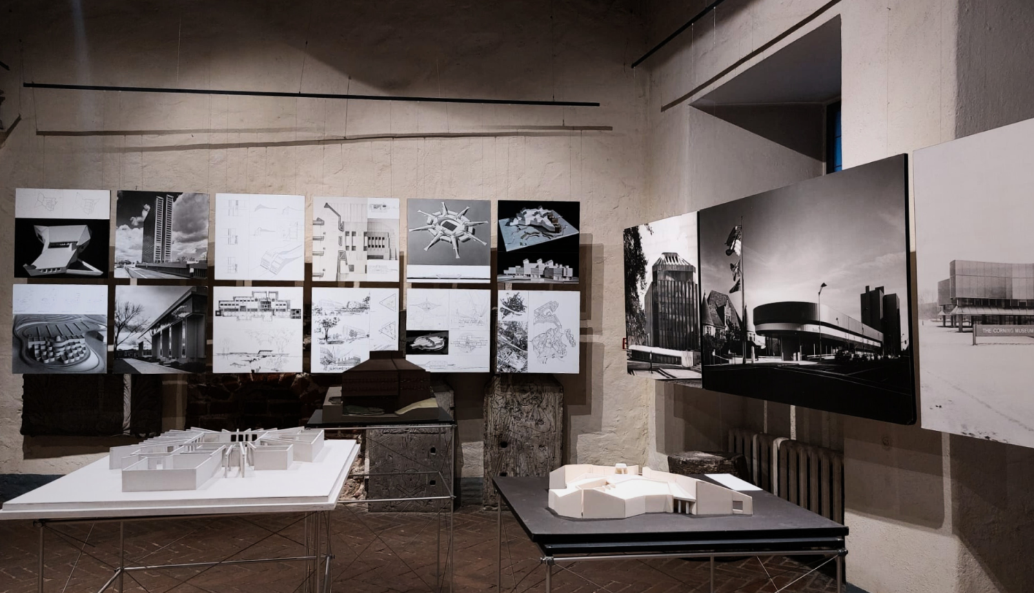 Fotoattēls no izstādes “Birkerts – 20. gadsimta akcenti” iekārtojuma Latvijas Arhitektūras muzejā. Telpā ar vēsturiskām, gaišām mūra sienām un tumšu ķieģeļu grīdu izvietoti arhitekta Gunāra Birkerta darbu eksponāti. Fonā pie sienas divās rindās piestiprinātas kvadrātveida planšetes ar melnbaltām ēku fotogrāfijām, skicēm un rasējumiem. Labajā pusē redzami trīs lielformāta vertikāli paneļi ar ikoniskām arhitektūras fotogrāfijām. Priekšplānā uz metāla karkasa galdiem novietoti divi arhitektūras maketi.