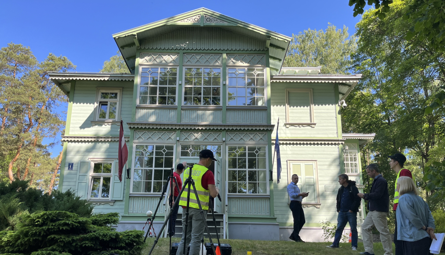 Attēlā redzama vēsturiska, gaiši zaļa koka ēka ar baltiem logu rāmjiem un dekoratīviem kokgriezumiem. Pie fasādes plīvo Latvijas karogs. Ēkas priekšā, zālienā notiek 3D skenēšanas darbi – vairāki cilvēki, daži no tiem dzeltenās atstarojošās vestēs, strādā ar mērniecības aprīkojumu. Kāds vīrietis gaišā kreklā aktīvi skaidro procesu pārējiem, kuri viņu vēro un sarunājas. Apkārt aug zaļi koki, un saulainais laiks rada patīkamu, vasarīgu atmosfēru.