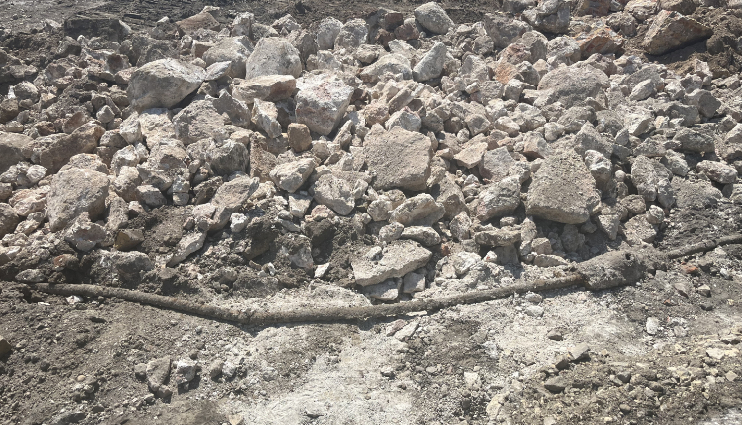 Būvlaukums ar izrakumiem un gružiem – priekšplānā redzami dažāda izmēra akmeņi un betona gabali, zem tiem daļēji atsegta gara, tumša caurule vai koka stienis. Fons ir klāts ar sausu, sablietētu zemi un tehnikas atstātām pēdām.