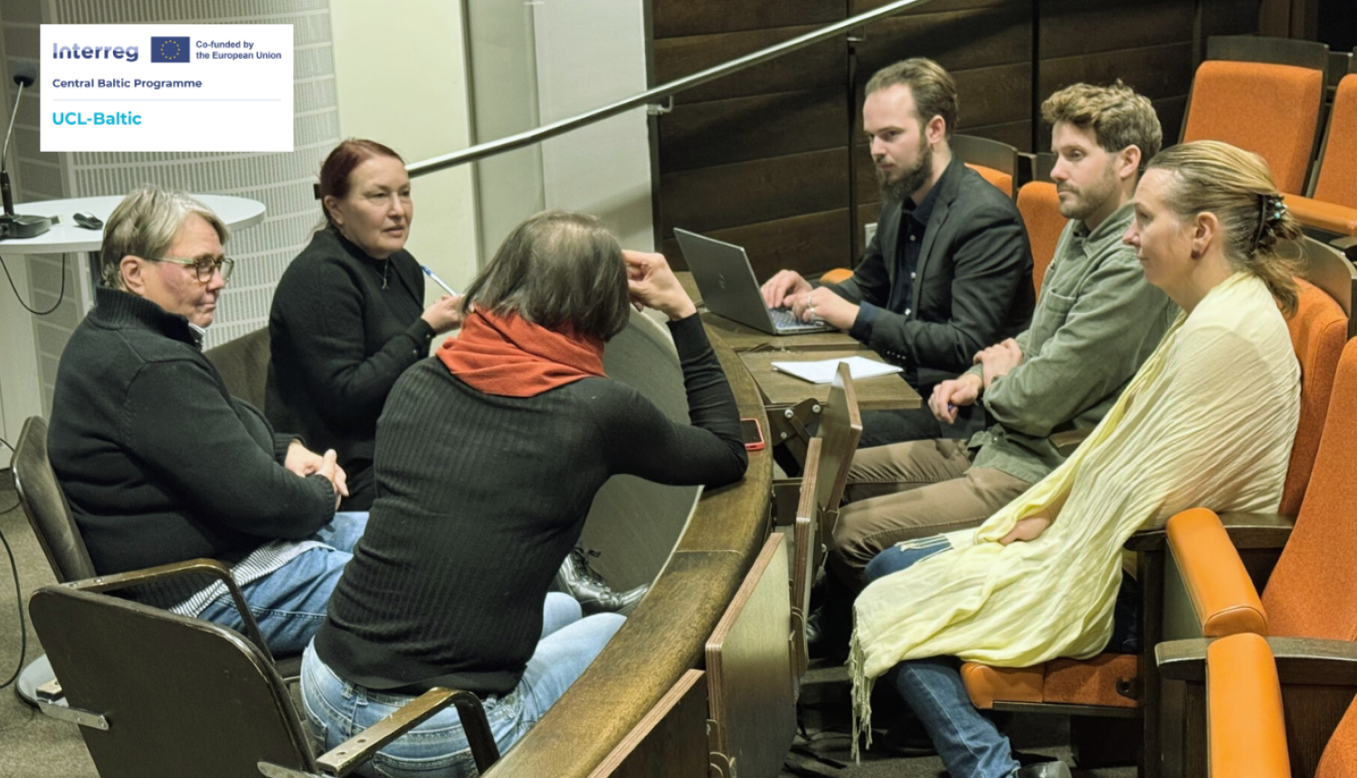 Semināra zālē seši projekta “UCL-Baltic” dalībnieki sēž ap apaļu galdu un pārrunā zemūdens kultūrvēsturisko ainavu izpētes jautājumus. Grupā ir trīs sievietes un trīs vīrieši, viens no dalībniekiem strādā pie klēpjdatora. Apkārt redzamas auditorijas rindas ar oranžiem krēsliem. Kreisajā augšējā stūrī ir projekta logo.