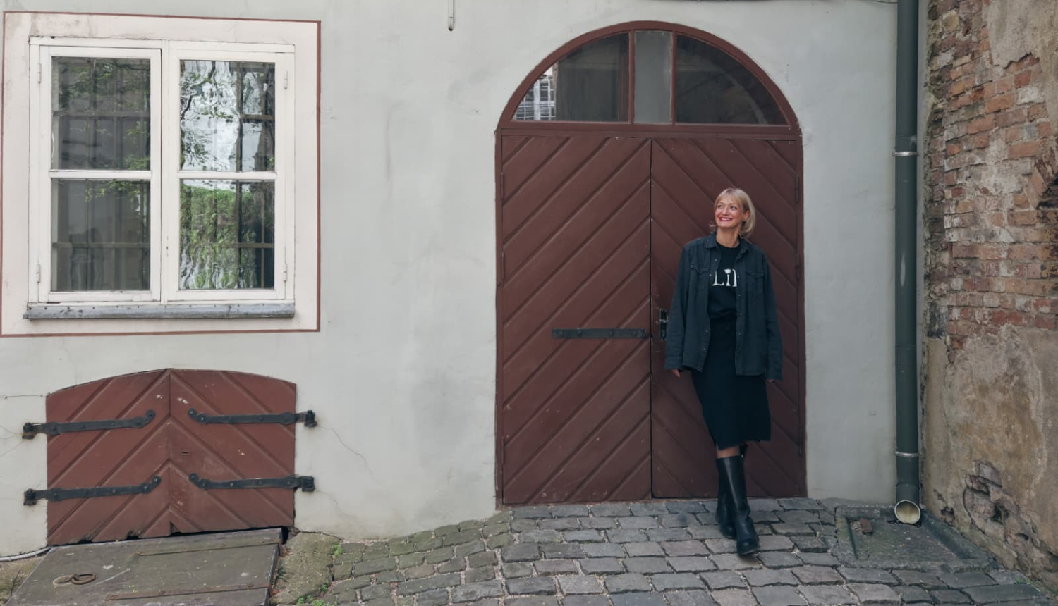 Pie vēsturiskas ēkas koka durvīm stāv gaišmataina sieviete tumšās drēbēs. Pa kreisi redzams neliels četru rūšu logs un pagraba lūka, pa labi - mūra siena. Zemi klāj bruģis.