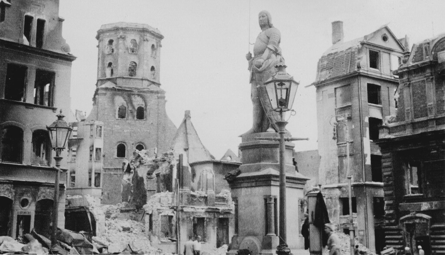 Melnbalta fotogrāfija, kurā redzams Otrā pasaules kara postījumu skats Rīgas Rātslaukumā. Attēlā redzama sagrauta pilsētas ainava ar gruvešiem un bojātām ēkām. Centrā izceļas Svētā Pētera baznīcas sagrautais tornis ar atklātām konstrukcijām, bet priekšplānā, starp iznīcinātām ēku fasādēm. Necietusi redzama Rolanda statuja. Attēls uzskatāmi demonstrē plašus pilsētas iznīcināšanas apjomus.