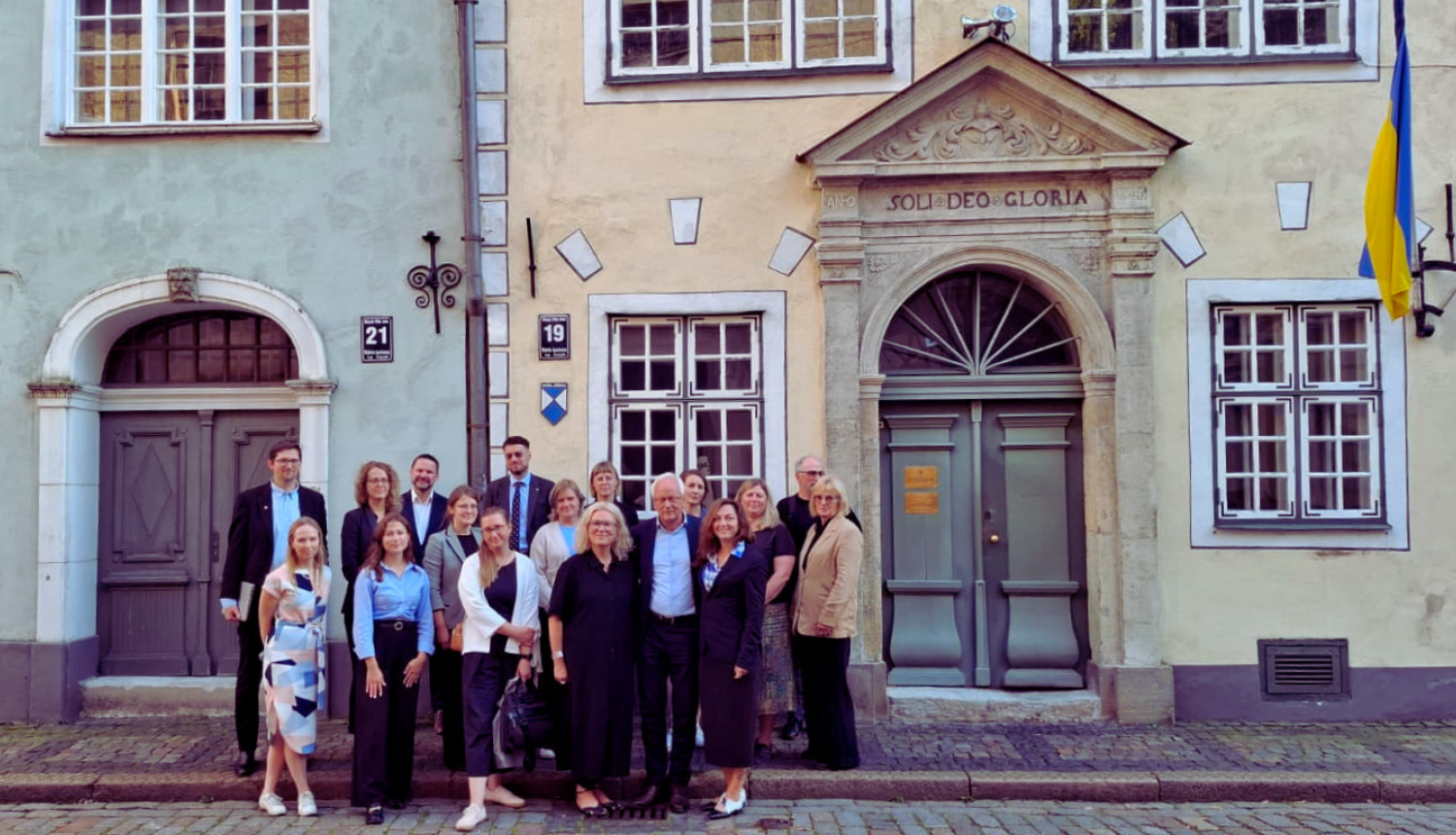 Attēls uzņems siltā dienā ārā, vēsturiskā pilsētvidē. Oficiālās drēbēs tērpta cilvēku grupa stāv uz vēsturiskā bruģa divu viduslaiku ēku priekšā. Grupā ir vairāk nekā 10 cilvēki, gan vīrieši, gan sievietes. Grupa atrodas attēla centrā, lejasdaļā. Fonā esošās ēkas ir zaļā un dzeltenā krāsā, tām ir sīkrūtiņu logi un viduslaiku ieejas portāli. Ēkai pa labi pašā sānā redzams piekārts Ukrainas karogs.