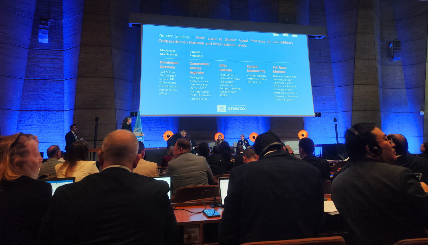 Plašā iekštelpu skatā redzama auditorija, kas sēž zālē un skatās uz lielu projekcijas ekrānu. Ekrānā ir balts teksts uz zila fona, kurā angliski norādīta plenārsesija: "No vietējā uz globālo: civilmilitārās sadarbības labā prakse nacionālā un starptautiskā līmenī". Zem virsraksta ir uzskaitīti runātāji un viņu tituli vai organizācijas. Apakšā labajā pusē ir redzams UNESCO logotips. Auditorija vērsta pret ekrānu, var redzēt daudzu cilvēku galvas un plecus. Tie sēž pie galdiem un klausās prezentāciju.  