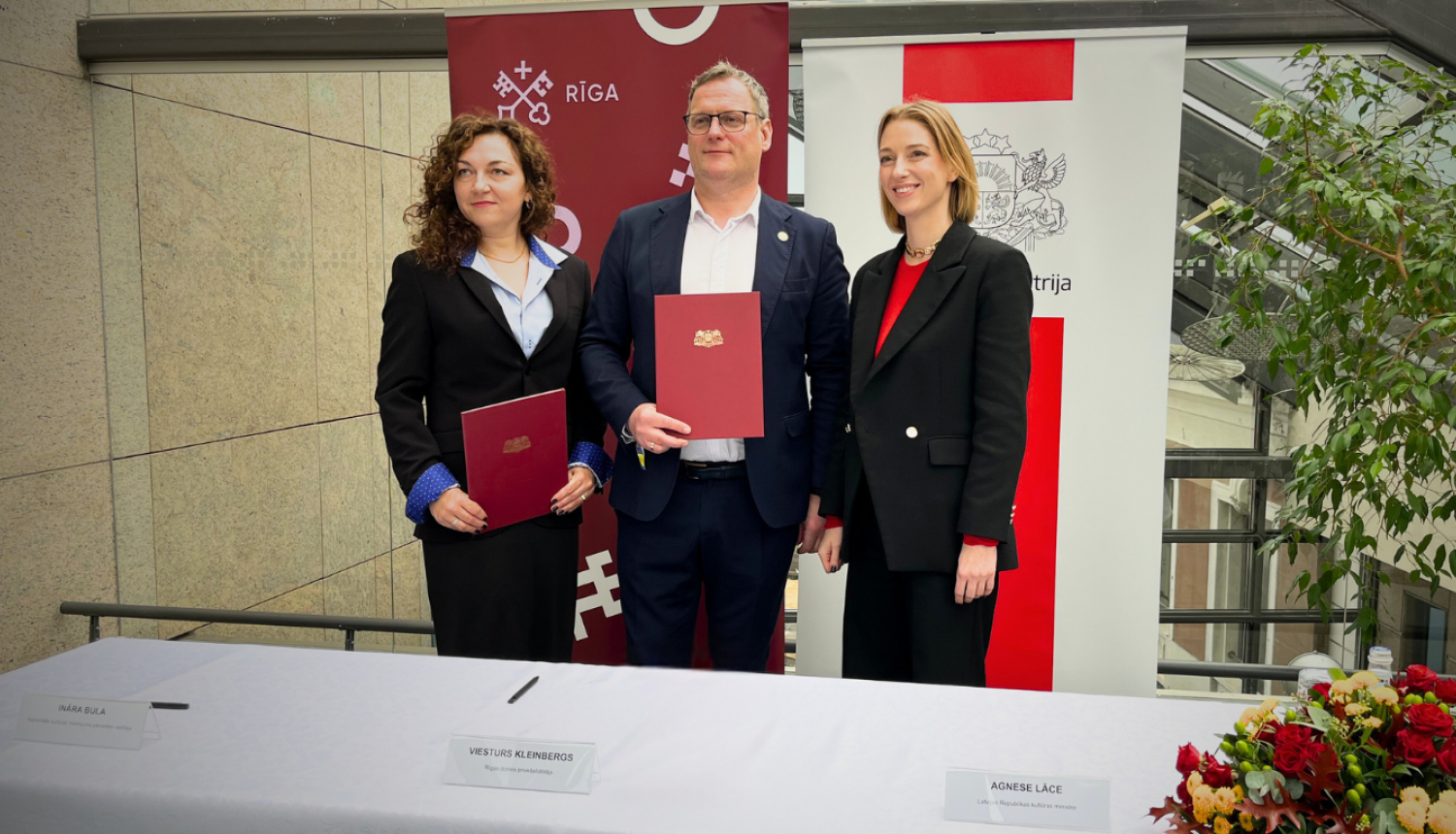 Priekšplānā atrodas galds, tas klāts ar baltu galdautu. Labajā apakšējā stūrī atrodas ziedu kārtojums, bet uz galda izkārtotas vārdu zīmītes. No labās puses - Ināra Bula, Viesturs Kleinbergs, Agnese Lāce. Visas trīs minētās personas stāv aiz galda un pozē bildei. Visi saģērbušies melnās žaketēs. Divi no cilvēkiem tur sarkanas mapes ar zeltītu Rīgas ģerboni. Aizmugurē redzami Rīgas pašvaldības un Kultūras ministrijas baneri un stikla siena. 