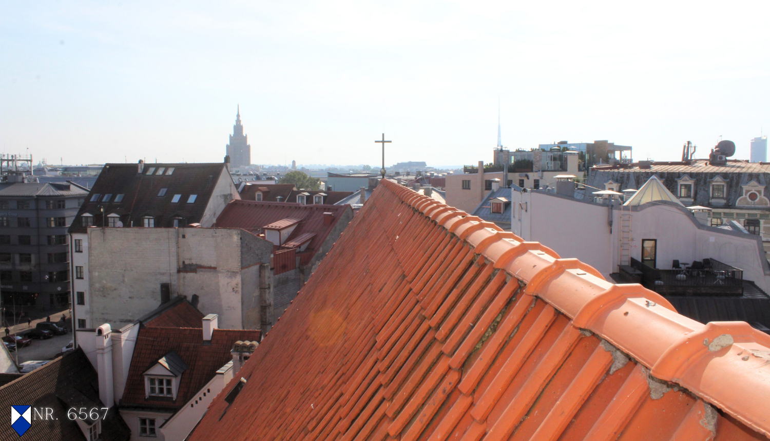 Attēlā redzams Rīgas Svētā Jāņa baznīcas jumta kores fragments tuvplānā. Jumts klāts ar sarkaniem dakstiņiem, kas veido izteiksmīgu tekstūru ar paralēlām līnijām. Jumta kores līnija stiepjas no attēla apakšējās labās puses uz augšējo kreiso pusi. Kores galā, attēla centrālajā daļā, pret debesīm paceļas tumšs metāla krusts. Fonā paveras plašs Vecrīgas panorāmas skats ar dažādu ēku jumtiem, kas ir galvenokārt sarkanos un pelēkos toņos. Fonā izceļas augsta, gaiša ēka ar smailu torni – Rīgas Zinātņu akadēmija. 