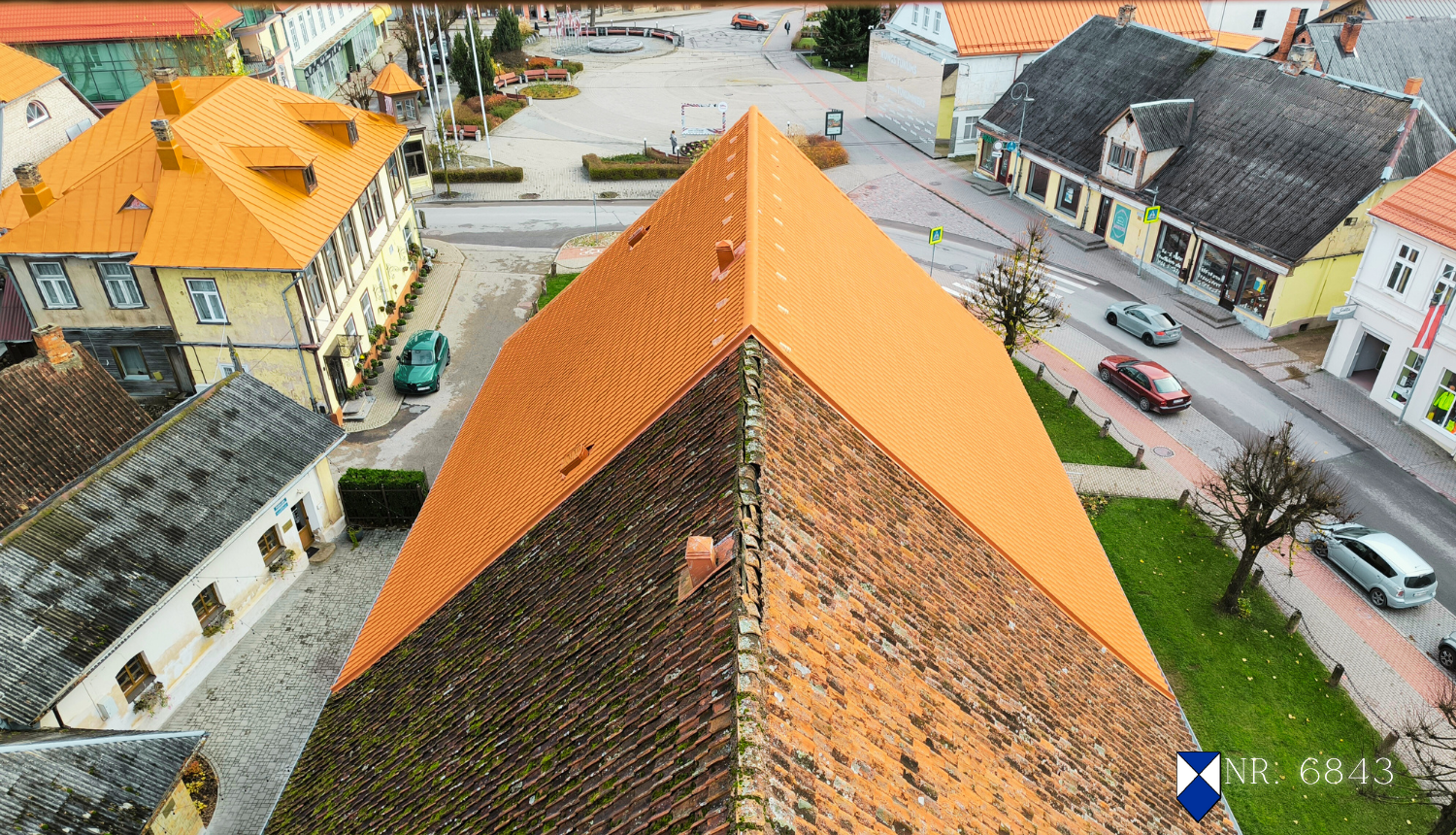 Skats no augšas uz Tukuma luterāņu baznīcas jumtu, kura viena puse nesen atjaunota ar spilgti oranžiem dakstiņiem, bet otra puse – vecāka, tumšāka, ar sūnām apaugusi. Baznīca atrodas pilsētas centrā, apkārt redzamas vēsturiskas ēkas ar dažādu veidu jumtiem, iela ar dažiem automobiļiem, gājēju ietves un koki. Attēla labajā apakšstūrī redzams kultūras pieminekļa simbols zili baltā krāsā un objekta numurs – 6843.