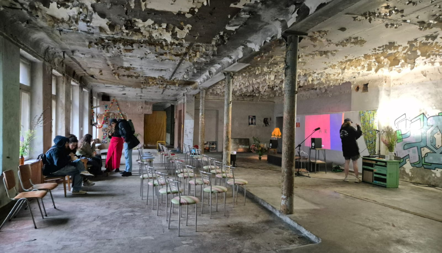 Attēlā redzami studenti. Tie atrodas plašā, neapdzīvotā vēsturiskā telpā. Tai no sienām un griestiem lobās krāsa, izskatās, ka tā tiek izmantota pagaidu pasākumiem. Grīda ir betona. Telpas vidū ir 15-20 metāla krēsli, kas vērsti ar muguru pret logiem. Telpas dziļumā ir neliela skatuve ar 2 mikrofoniem un projektoru, kas projicē rojicē gaiši rozā-purpursarkanu attēlu uz baltas sienas. Skatuves labajā pusē ir grafiti uzraksts, un uz grīdas ir novietotas puķu vāzes. 