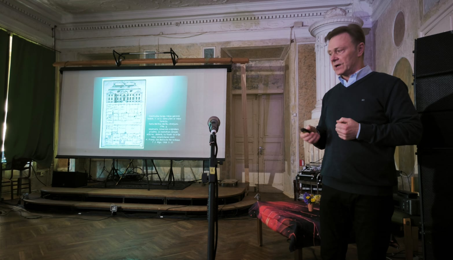 Skats ar prezentāciju. Kreisajā pusē stāv tumšās drēbēs ģērbies vīrietis ar īsiem matiem. Blakus atrodas mikrofons. Dziļumā redzama uz izvelkama ekrāna projicēta prezentācija. Viss notiek vēsturiskā telpā.