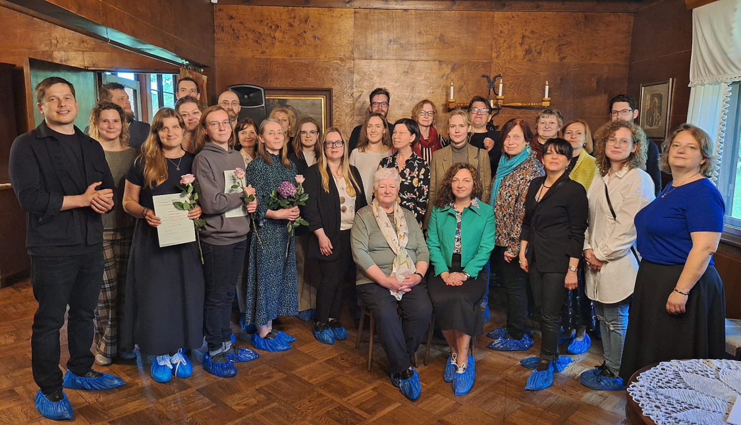 Grupa aptuveni 25-30 cilvēku, vīrieši un sievietes, daži ar apliecībām rokās un ziediem, stāv un sēž telpā ar koka apdari, vēsturiskā vidē. Visiem ir zili apavu pārvalki, lai pasargātu koka parketa grīdu. Attēls iemūžina restauratoru kvalifikācijas celšanas apliecību pasniegšanas pasākumu.