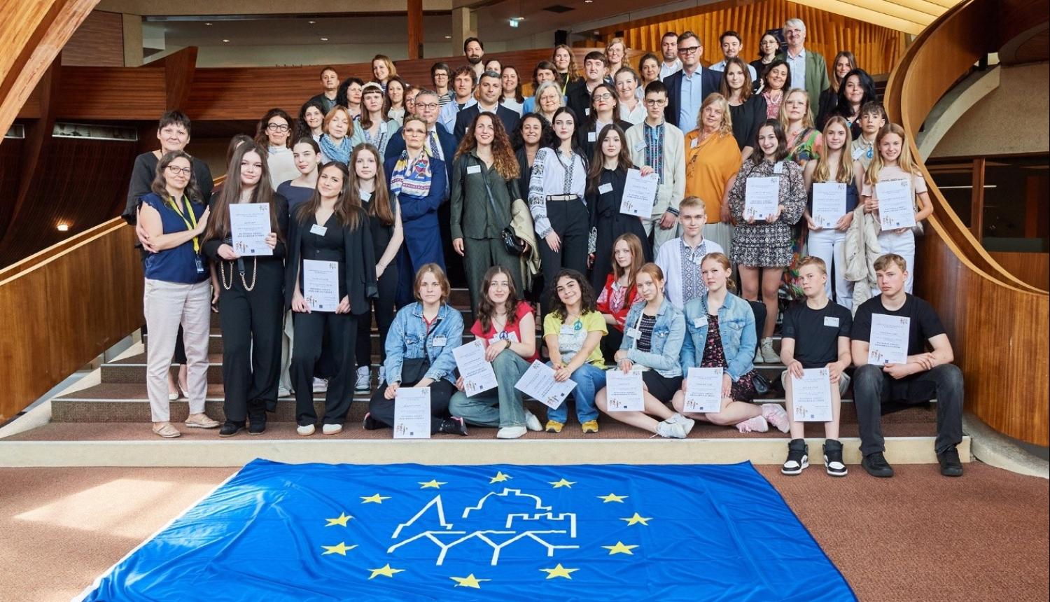 Smaidīga jauniešu un pieaugušo grupa stāv pozēdami attēlam, tiem priekšā izklāts Eiropas kultūras mantojuma dienu karogs. Karogs ir līdzīgs Eiropas savienības karogam, bet tam pa vidu ir ēku kontūras. Apkārt redzama plaša telpa ar koka interjeru.