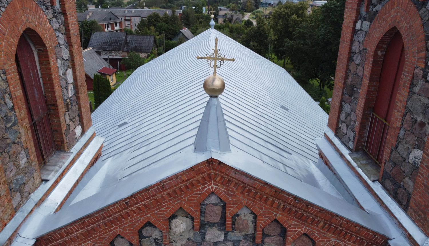 Attēlā redzams restaurētas baznīcas jumta fragments no putna lidojuma perspektīvas. Galvenajā plānā ir liels, lēzens divslīpju jumts, kas klāts ar gaiši pelēkām skārda loksnēm. Jumta korē pacelas metāla smaile ar zeltītu dekoratīvu lodi un krustu. Abās pusēs jumtam piekļaujas masīvi torņi, kas būvēti no sarkanajiem ķieģeļiem un neapstrādātiem laukakmeņiem, kuros redzamas arkveida logu ailes. Fonā pavīd nelielas ciemata mājas un koki, uzsverot objekta nozīmību vietējā ainavā.