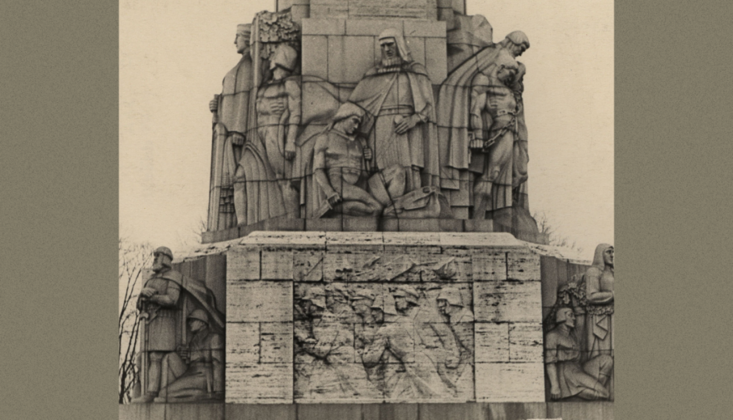 Attēlā uz pelēcīga fona redzama melnbalta fotogrāfija, kurā ir redzama Brīvības pieminekļa daļa, kas vērsta pret Operas pusi. Tajā izcelts cilnis, kas attēlo Bermontiādi, kas saistīts ar Brīvības cīņām un to, kāpēc svinam Lāčplēša dienu.