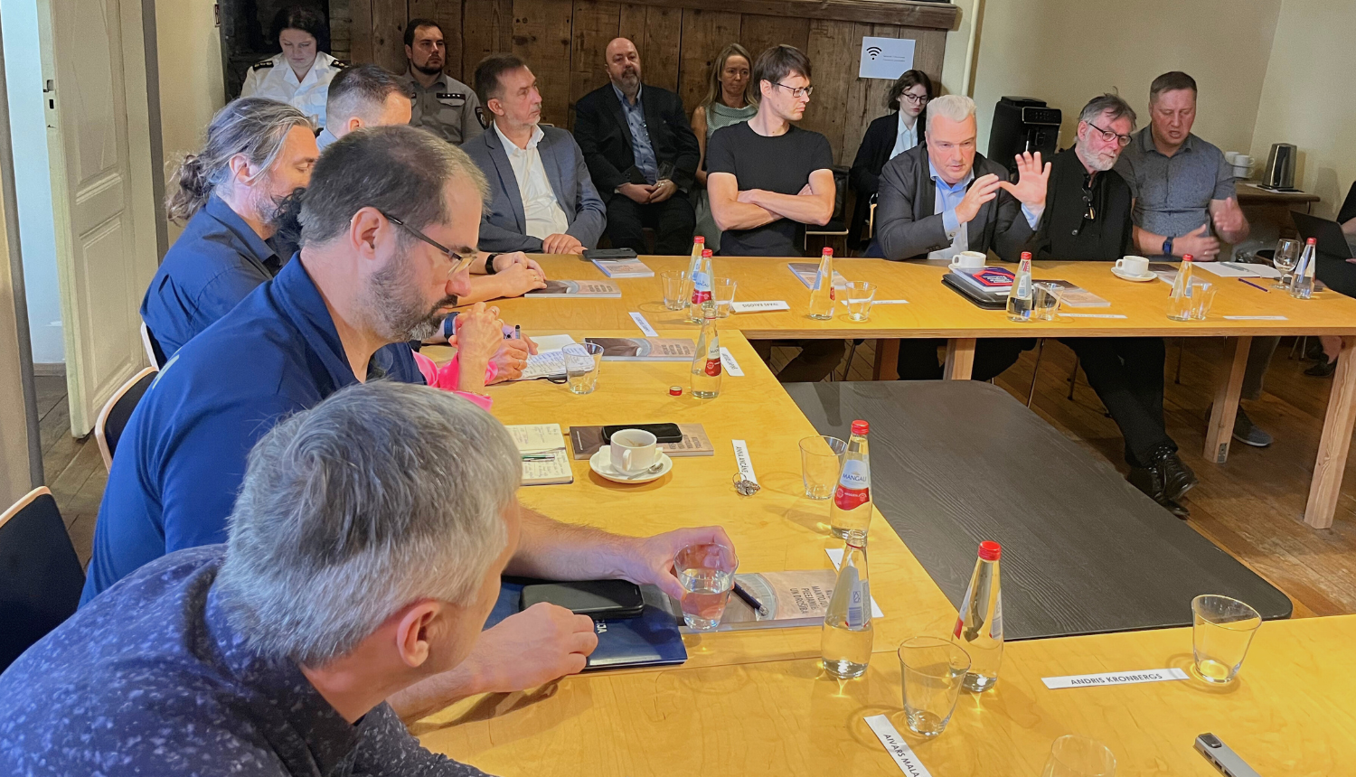 Nacionālajā kultūras mantojuma pārvaldē pie U veida galda sēž Apeirona, Valsts Policijas, Ugunsdzēsības un glābšanas dienesta, Latvijas Militāro spēku pārstāvji, arhitekti, Nacionālās kultūras mantojuma pārvaldes speciālisti, ģeotelpisko datu speciālists. Visi iegrimuši diskusijā.