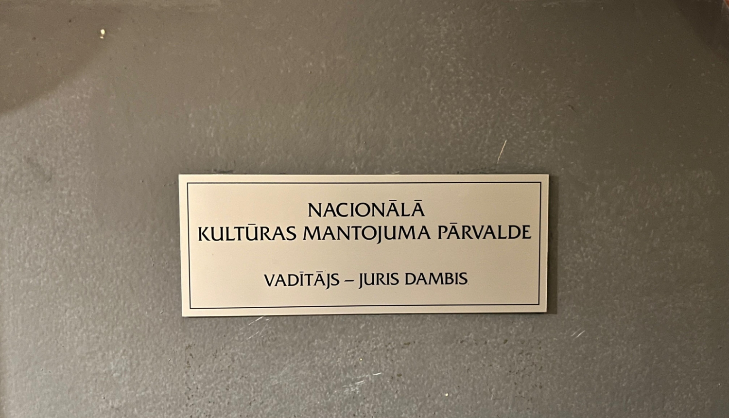 Plāksnīte pie Nacionālās kultūras mantojuma pārvaldes vadītāja durvīm, kas saka "Nacionālā kultūras mantojuma pārvalde"; vadītājs - Juris Dambis. Krāsotās koka durvis ir pelēcīgas, plāksne četrkantīga, neliela, metāla ar melniem burtiem.