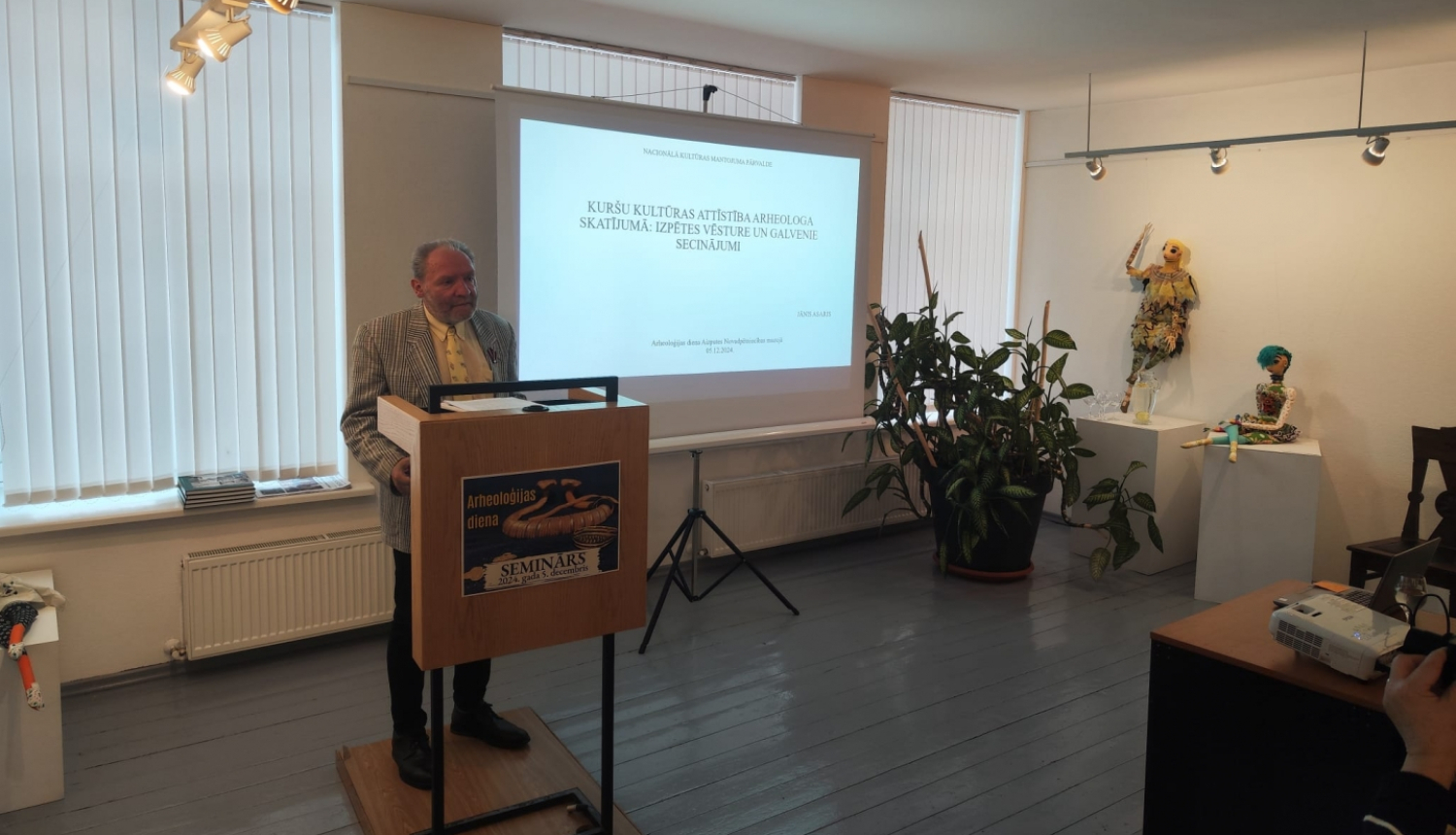 Gaišā telpā auditorijas priekšā stāv vīrietis ar sirmiem matiem, rūtotā uzvalkā. Vīrietis stāv pie pults un viņam aiz muguras ir ekrāns ar prezentāciju