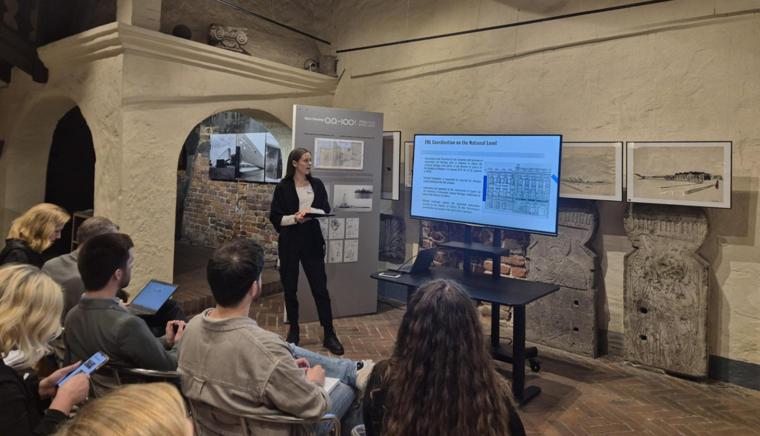 Attēlā redzama vēsturiskas ēkas telpa ar gaišām apmestām sienām, sarkanu ķieģeļu grīdu un brūnām griestu sijām. Kreisajā pusē fonā redzams apvalkdūmenis. Attēla lejasdaļā krēslos redzami cilvēki, kas klausās prezentāciju. Attēla vidū, dziļumā redzama sieviete oficiālā kostīmā, kas kaut ko stāsta un rāda uz digitālo ekrānu, kurā redzama prezentācija.