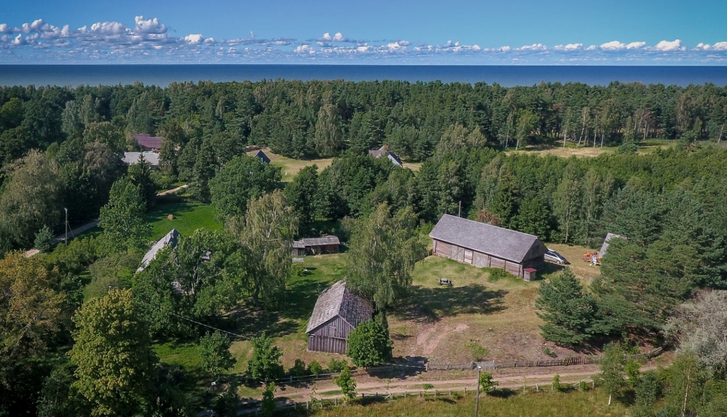 Attēlā redzams pilsētbūvniecības piemineklis - Košraga zvejniekciems. Pilsētbūvniecības piemineklis iekļauj plašāku cilvēka veidotu vidi, kā vecpilsētu vai ciemu. Attēlā redzams ar dronu no gaisa fotografētais Košraga ciems lībiešu krastā. Foto augšpusē pirmo ceturtdaļu aizņem tumši zilas vasaras debesis ar rotaļīgu baltu mākonīšu rindu virs horizonta, kam seko tumši zaļais priežu mežs. Priekšplānā var izšķirt kādu no ciema vēsturiskajām ēkām, starp kokiem šur tur manāmi zemi divslīpju jumti.