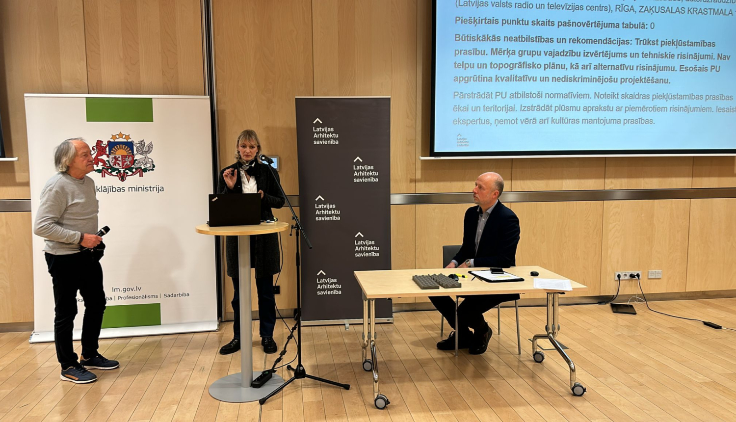 Attēlā redzama konferenču telpa koka interjerā. Telpā redzami 3 cilvēki. Pa kreisi stāv sirms kungs pelēkā džemperī un melnās biksēs. Pa vidu pie stāvgalda stāv gara sieviete ar gaišiem matiem tumšās drēbēs. Labajā pusē pie galda sēž kungs melnā uzvalkā. Aiz cilvēkiem ir Labklājības ministrijas un Arhitektu savienības baneri. Labajā sānā redzams ekrāns uz kura atspoguļojas teksts.